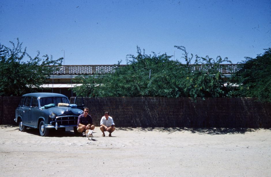 1950s scene with men, dog, and a blue car in a desert, showcasing vintage style.